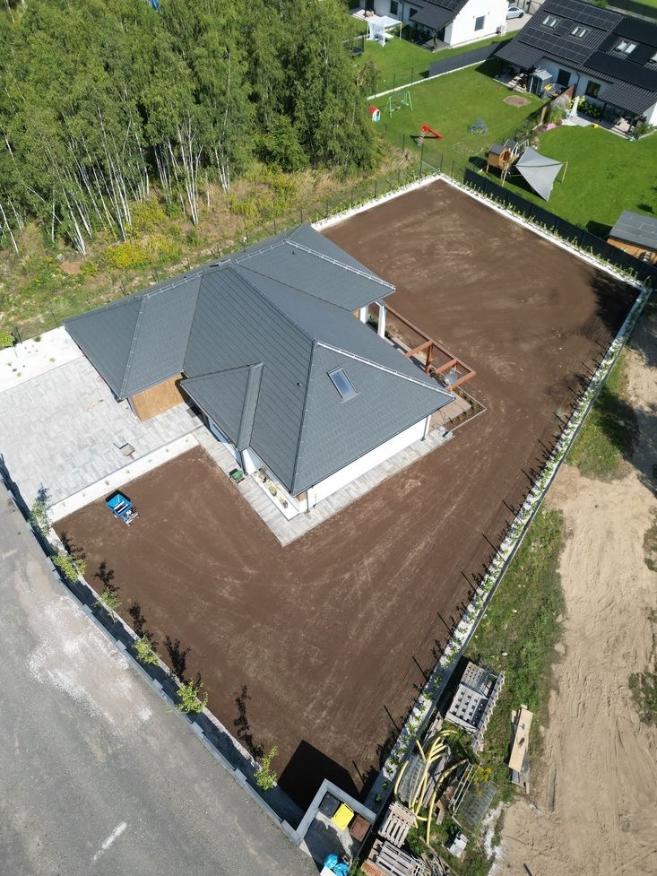 Widok z drona na przygotowany teren pod trawnik wokół domu z szarą dachówką i kostką brukową, z widoczną glebogryzarką i paletami z rurami nawadniającymi.