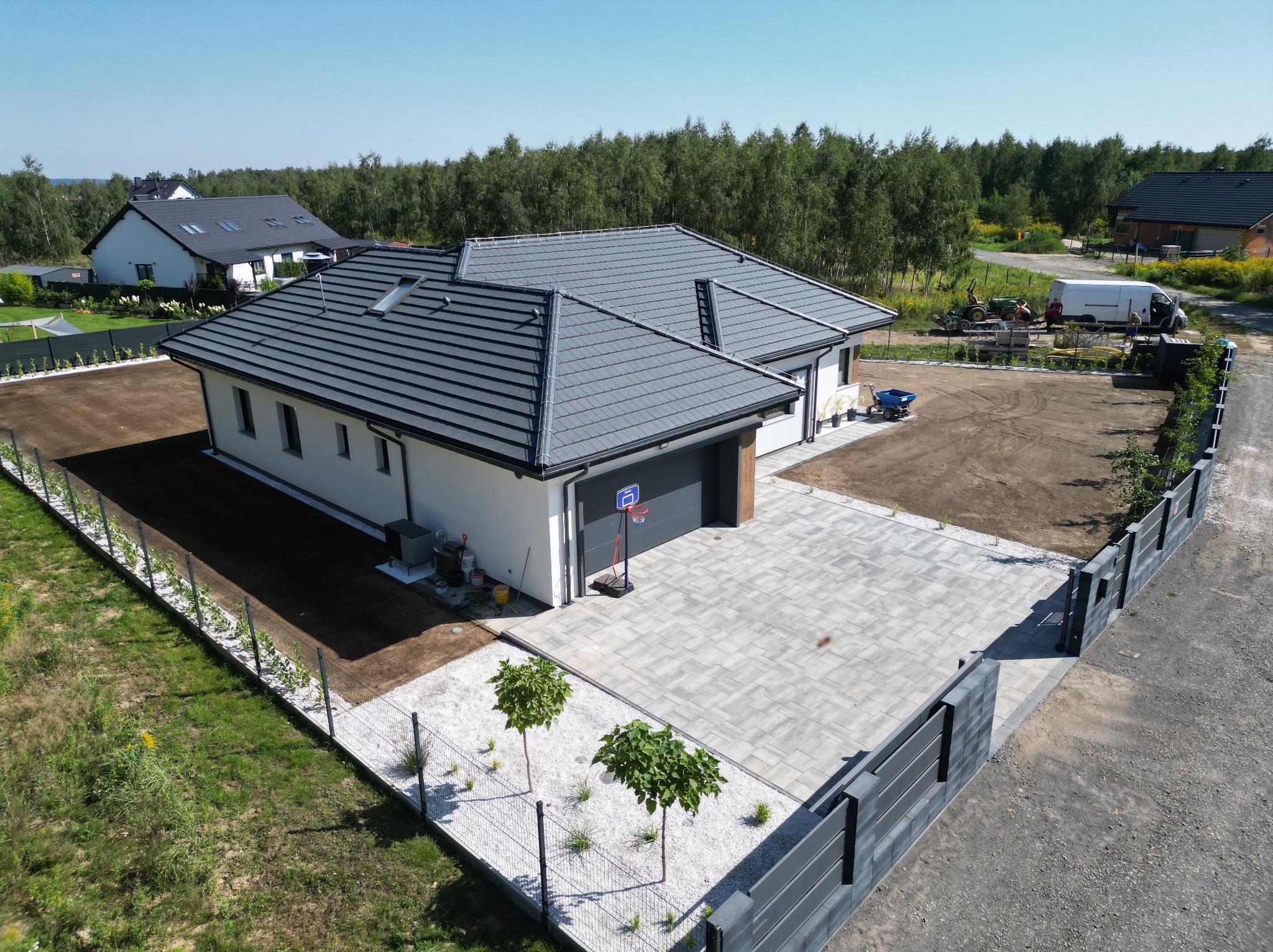 Widok z góry na posesję z nowo założonym trawnikiem, utwardzonym podjazdem z szarej kostki, ogrodzeniem panelowym i nasadzonymi młodymi drzewkami.