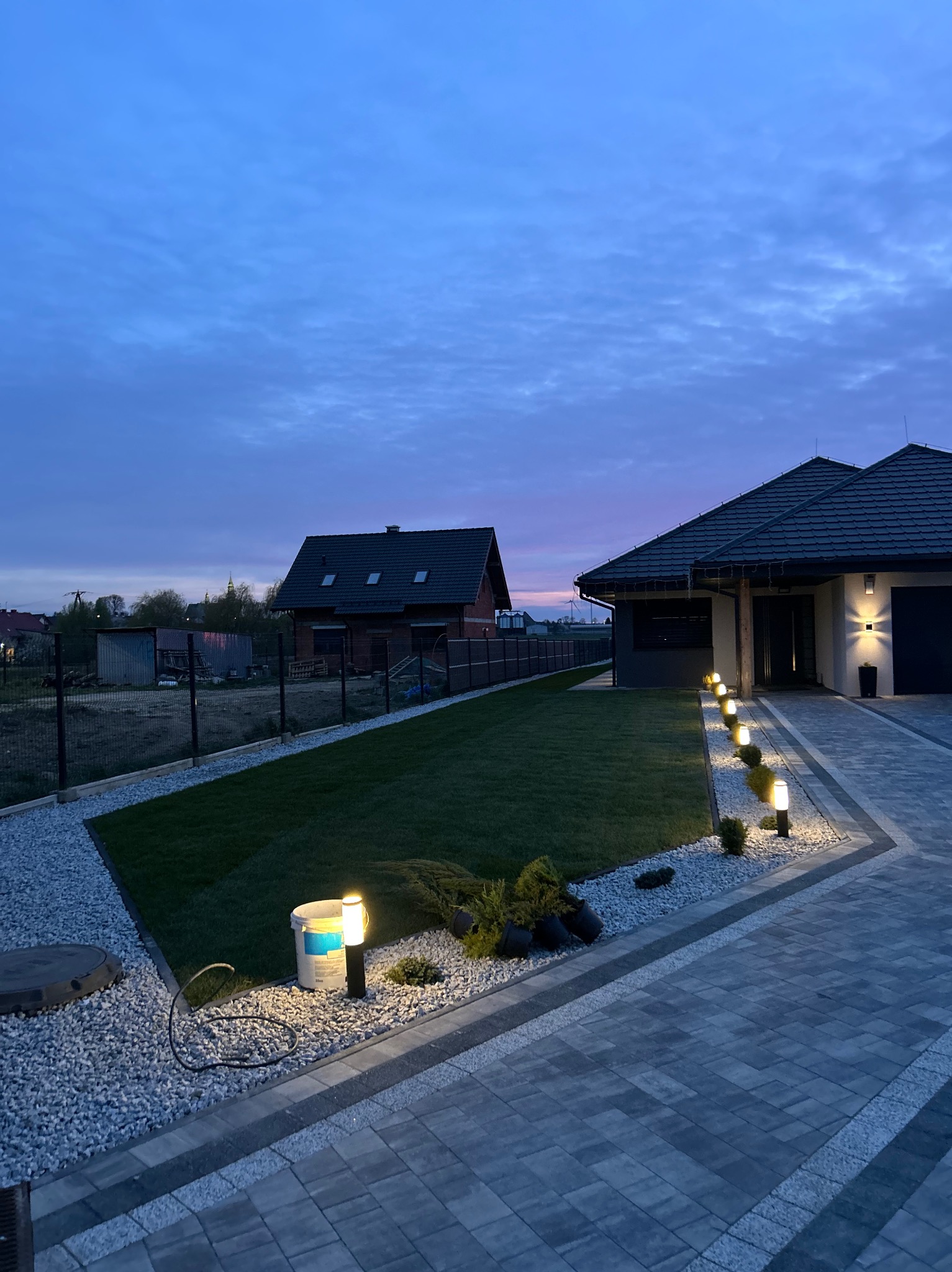 Oświetlona posesja z równo przystrzyżonym trawnikiem, ozdobnym kamieniem i ścieżką z kostki brukowej w odcieniach szarości. W tle widoczny dom i wieczorne niebo.
