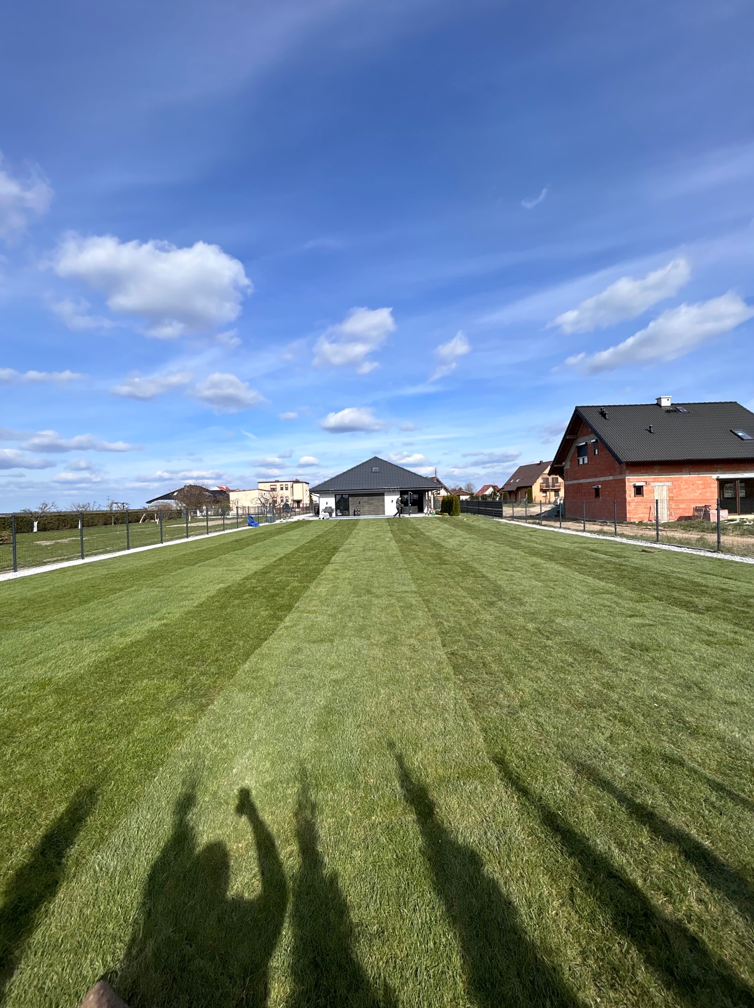 Rozległy trawnik z równo przystrzyżoną trawą w pasy, otoczony ogrodzeniem panelowym i zabudową jednorodzinną w tle, w słoneczny dzień.