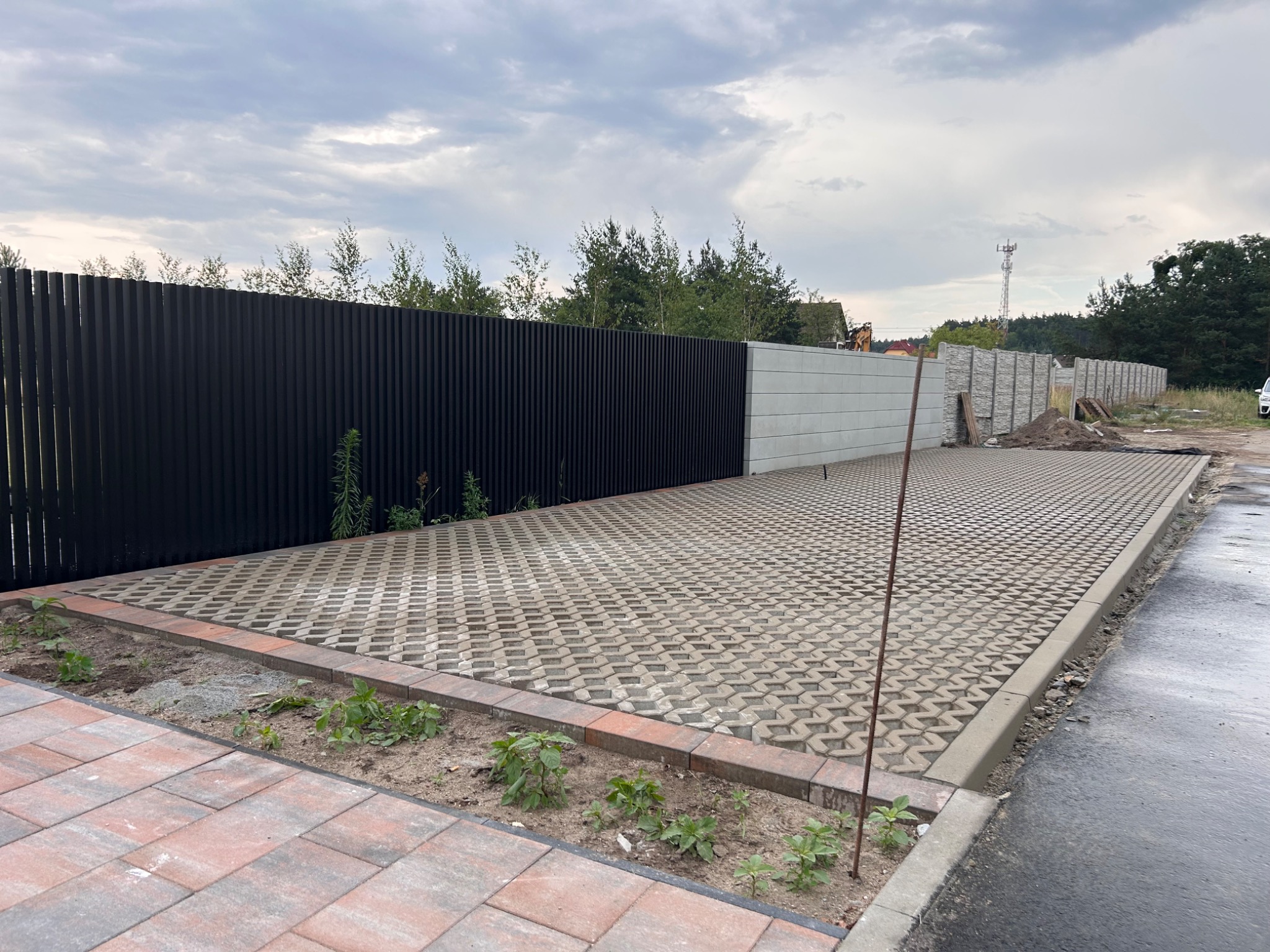 Ukończona nawierzchnia z ażurowych płyt betonowych na podjeździe, obrzeża z kostki, w tle nowoczesne ogrodzenie panelowe i betonowe, pochmurne niebo.