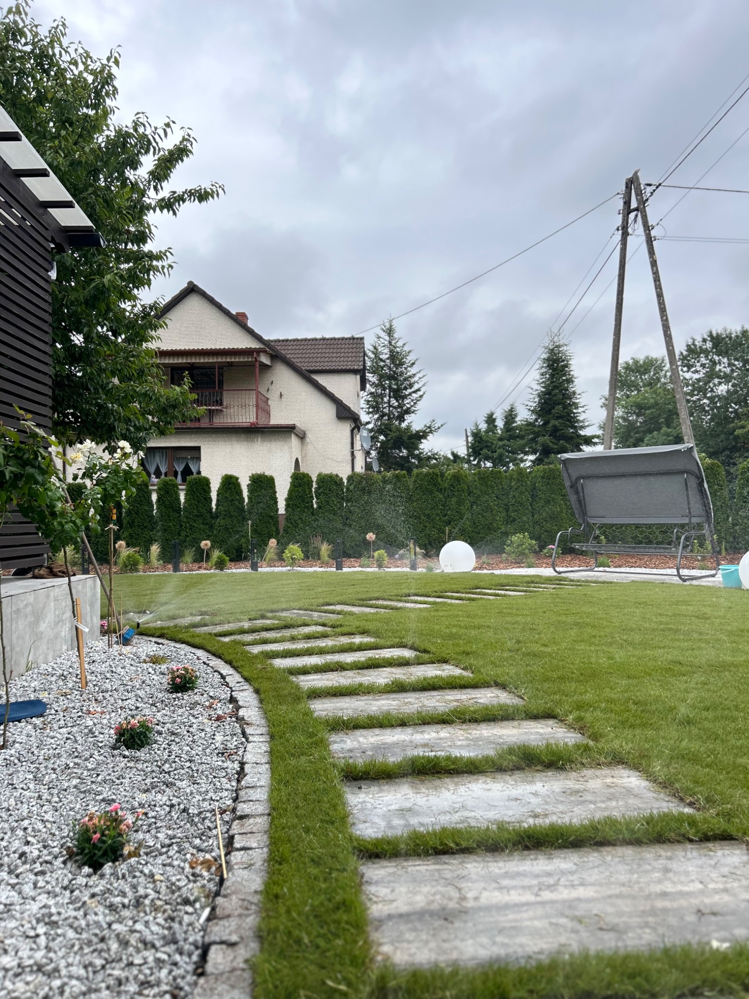 Nowoczesny ogród z trawnikiem, ścieżką z płyt, rabatami z kamienia, niskimi krzewami i systemem nawadniania w trakcie pracy. W tle dom i huśtawka.