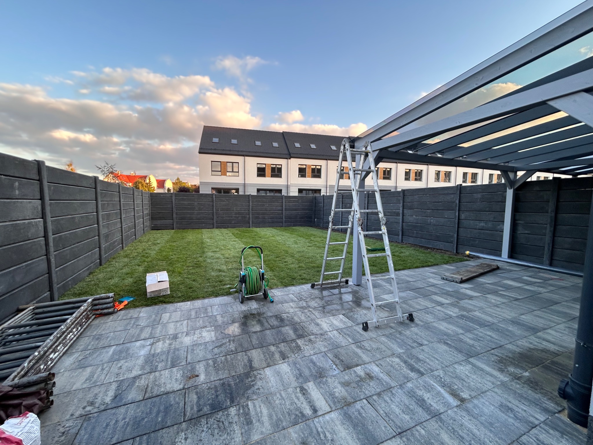 Świeżo ułożona trawa z rolki w ogrodzie, widok z tarasu z szarych płyt. Na trawniku wąż ogrodowy i drabina. Szare ogrodzenie panelowe w tle.