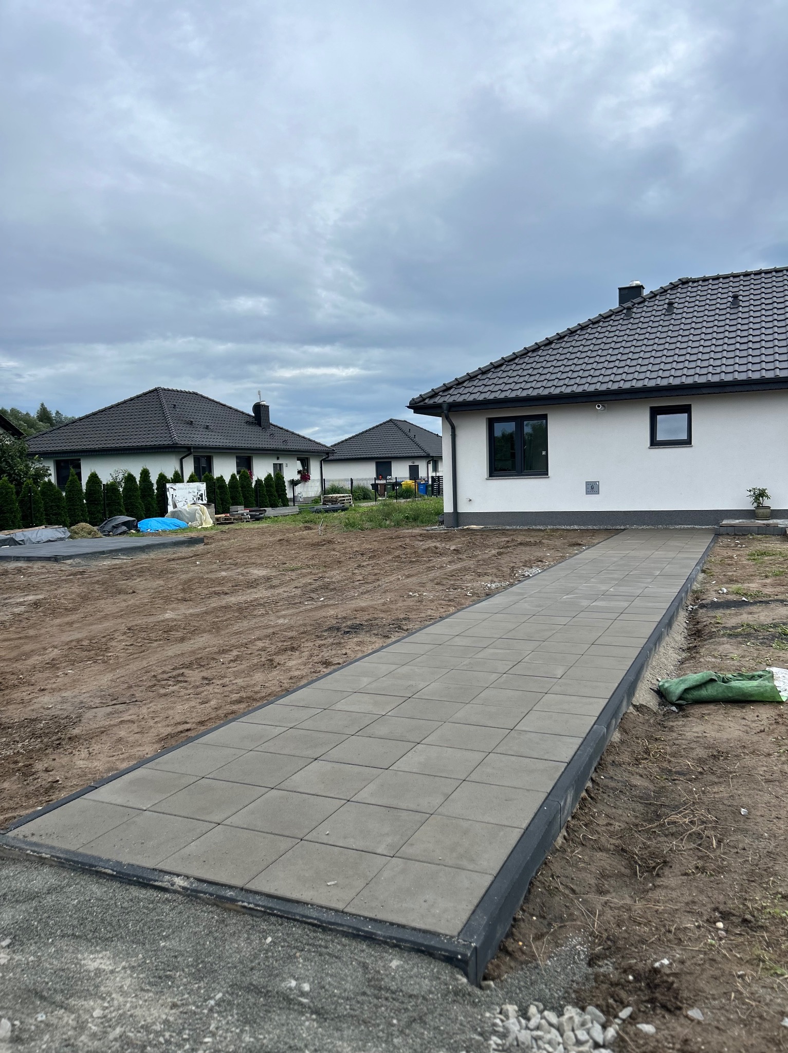 Ułożony chodnik z szarych kostek brukowych prowadzący do domu, otoczony ziemią przygotowaną pod trawnik. Widoczne domy w tle i pochmurne niebo.