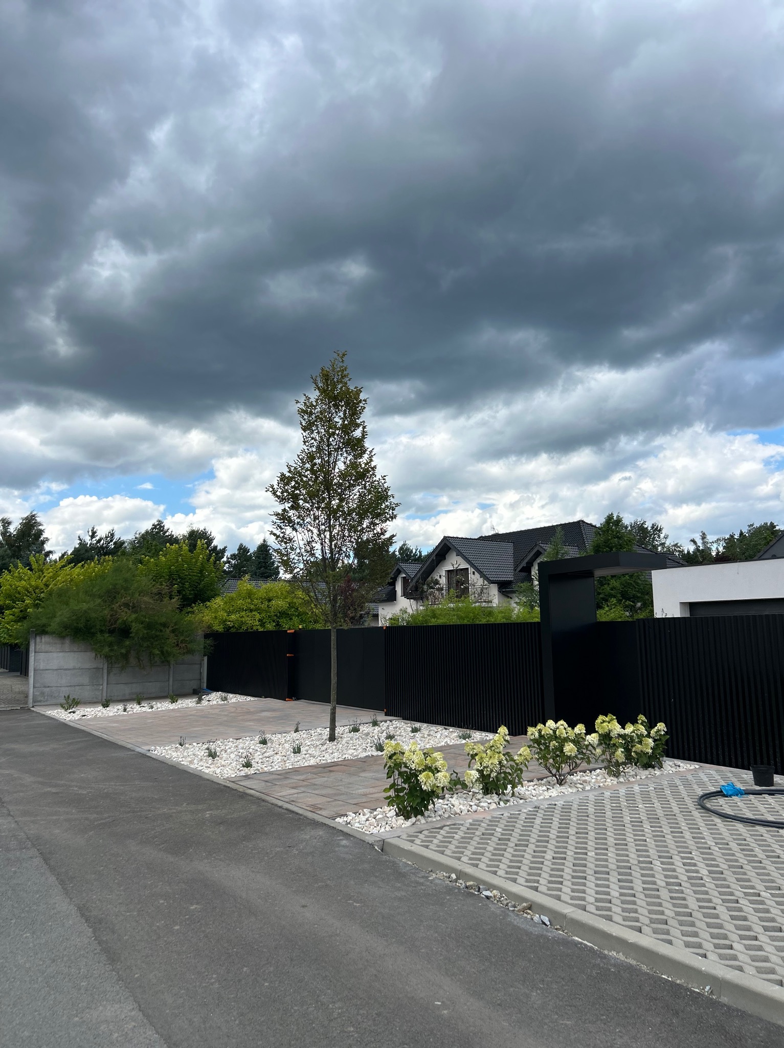 Nowoczesna aranżacja przestrzeni przed domem: kostka ażurowa, białe kamienie, rabaty z roślinami, drzewo i czarne, pionowe ogrodzenie na tle pochmurnego nieba.