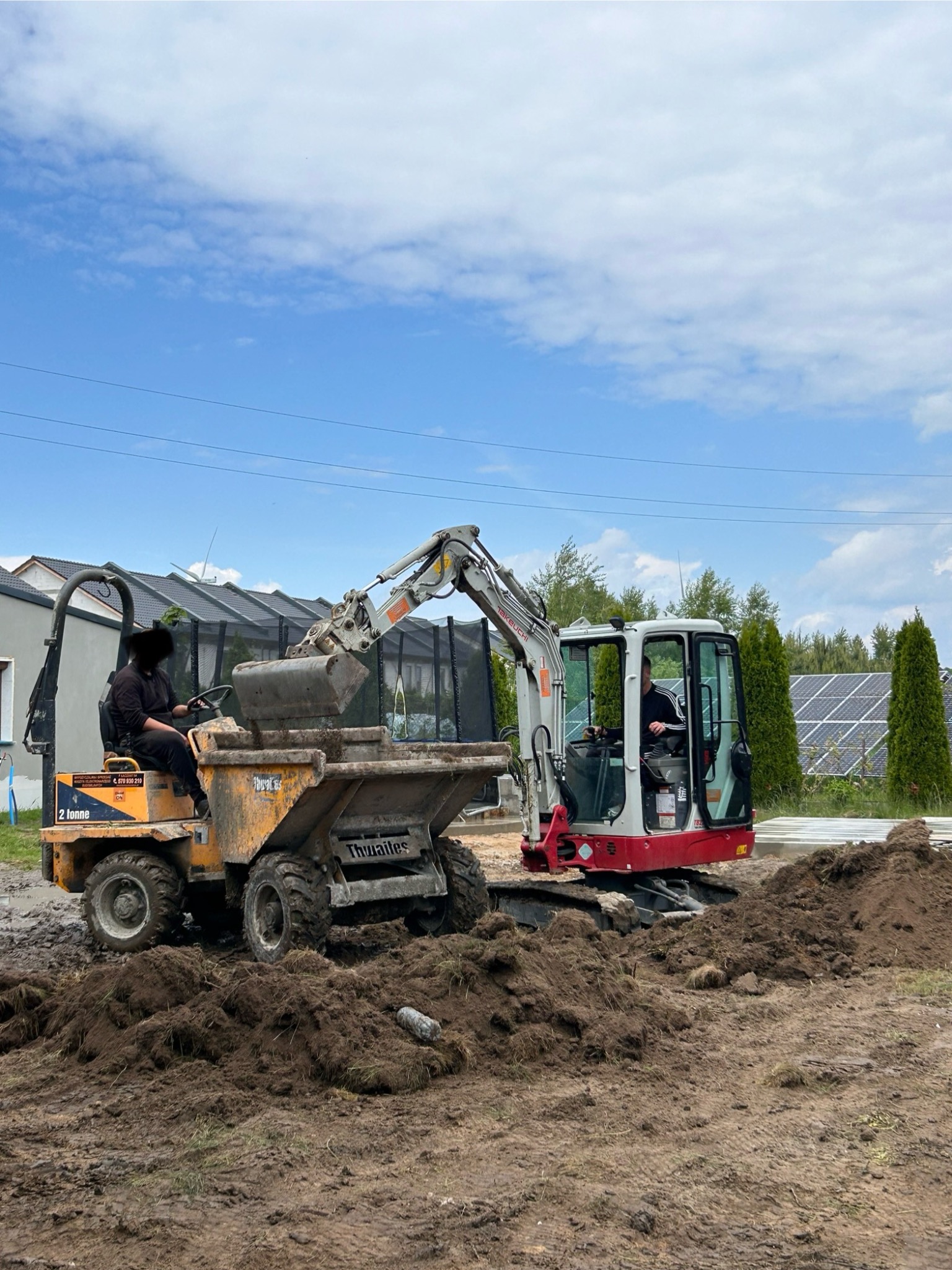 Żółta wywrotka i koparka na placu budowy. Operator koparki nabiera ziemię do wywrotki. W tle widoczne panele słoneczne i budynki mieszkalne.