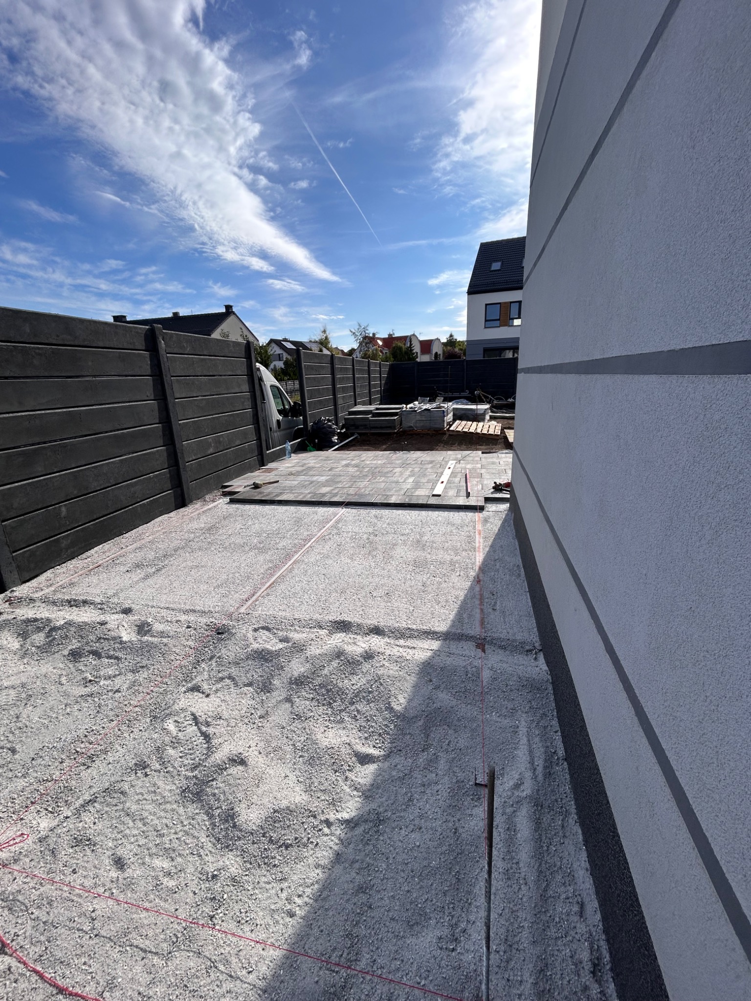 Etap układania kostki brukowej na podsypce z piasku, widoczne sznurki wyznaczające obszar. W tle paleta z kostką i fragment ogrodzenia z desek kompozytowych.