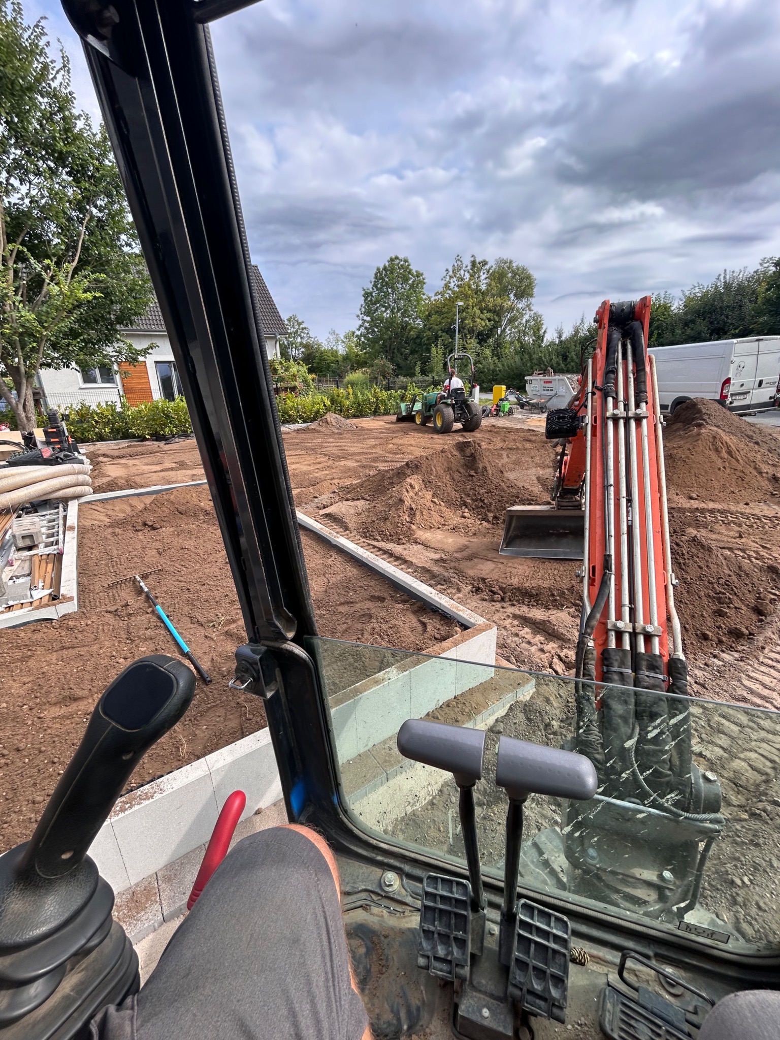 Widok z kabiny koparki na plac budowy: wykop pod fundamenty, przygotowany teren, sprzęt budowlany. Perspektywa operatora maszyny.