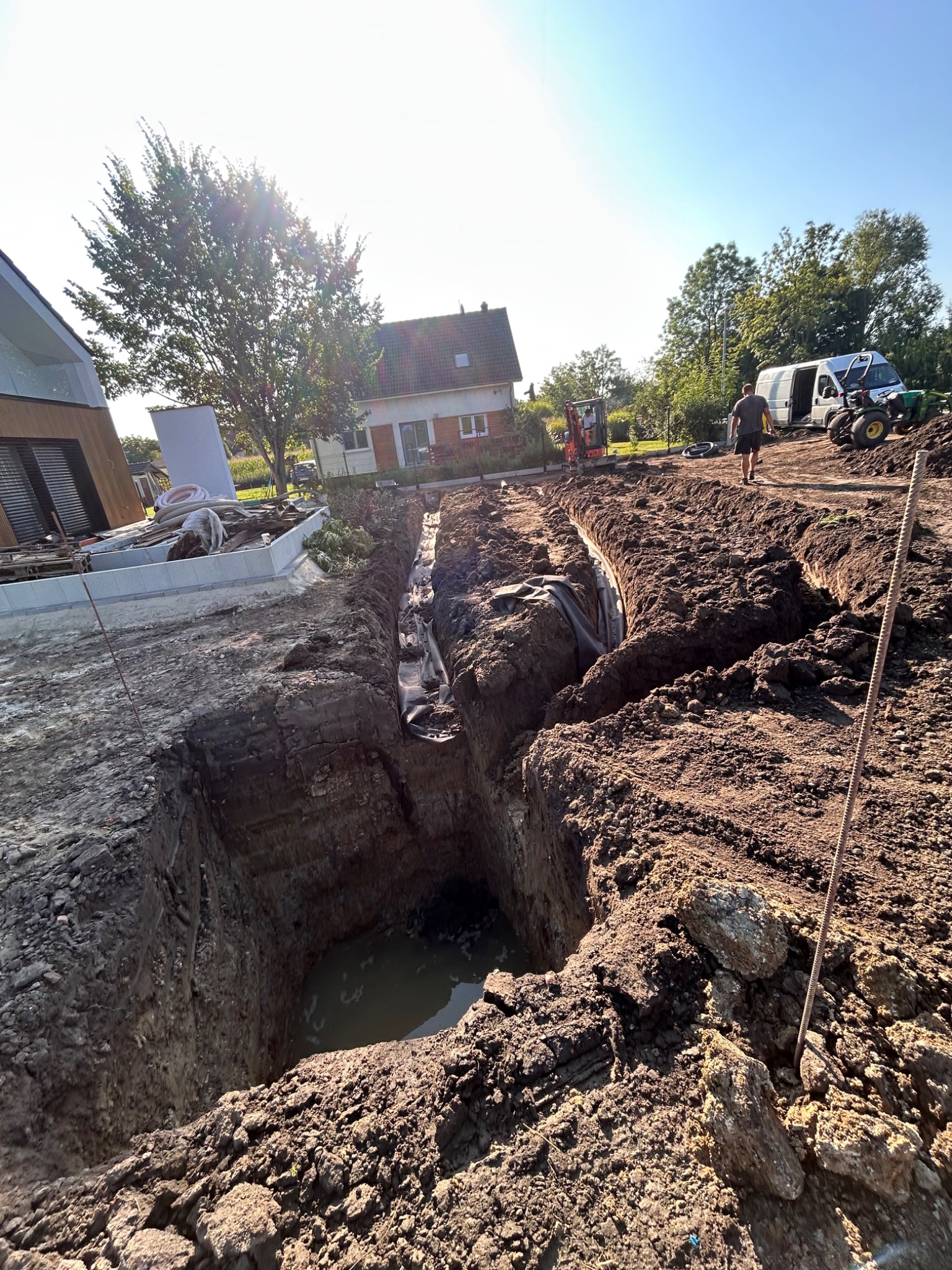 Głęboki wykop w ziemi z wodą na dnie, widoczne rury i folia, w tle budynki, drzewa, sprzęt budowlany i osoba. Słoneczny dzień, niebieskie niebo.