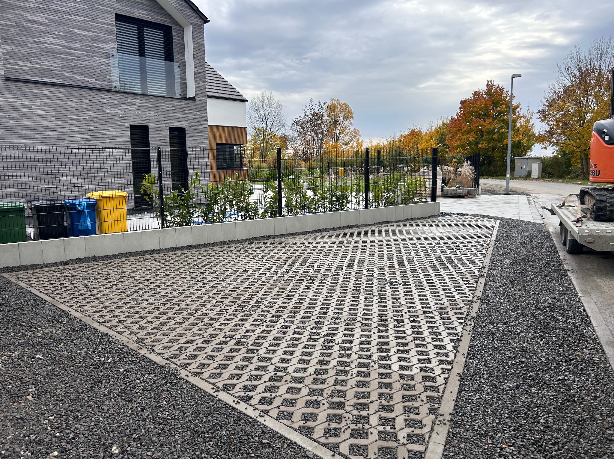 Szara ażurowa nawierzchnia z wypełnieniem żwirowym na posesji, obok nowoczesny dom z szarej cegły i ogrodzenie z siatki. Widoczny fragment maszyny budowlanej.