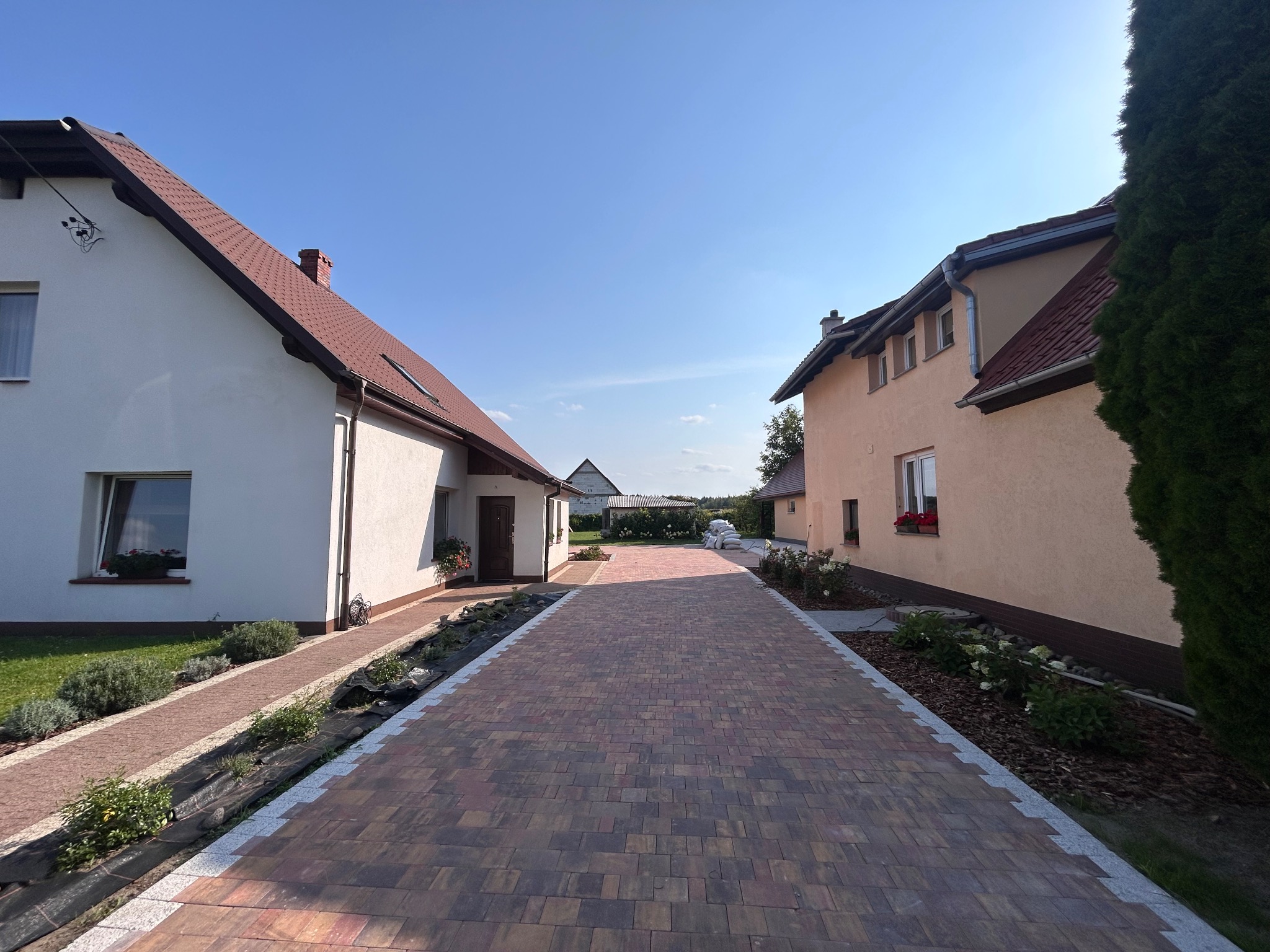 Ułożona kostka brukowa tworzy ścieżkę między dwoma domami. Widoczne rabaty z roślinami i trawnik. Słoneczny dzień, błękitne niebo. Kostka w odcieniach brązu i czerwieni.