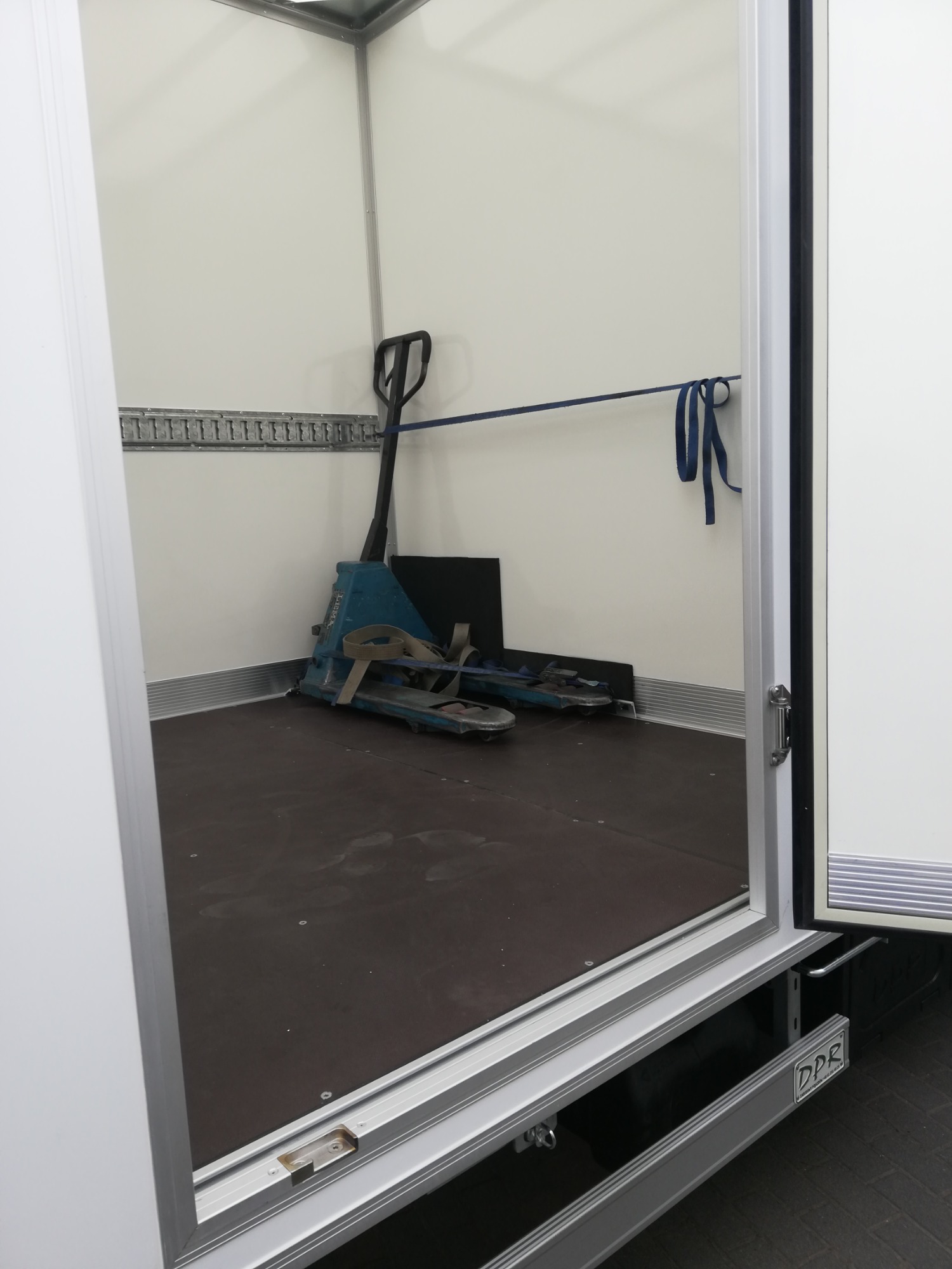 Wnętrze pustej, białej furgonetki z brązową podłogą, widoczny wózek paletowy w rogu, zabezpieczony niebieskim pasem do ściany.