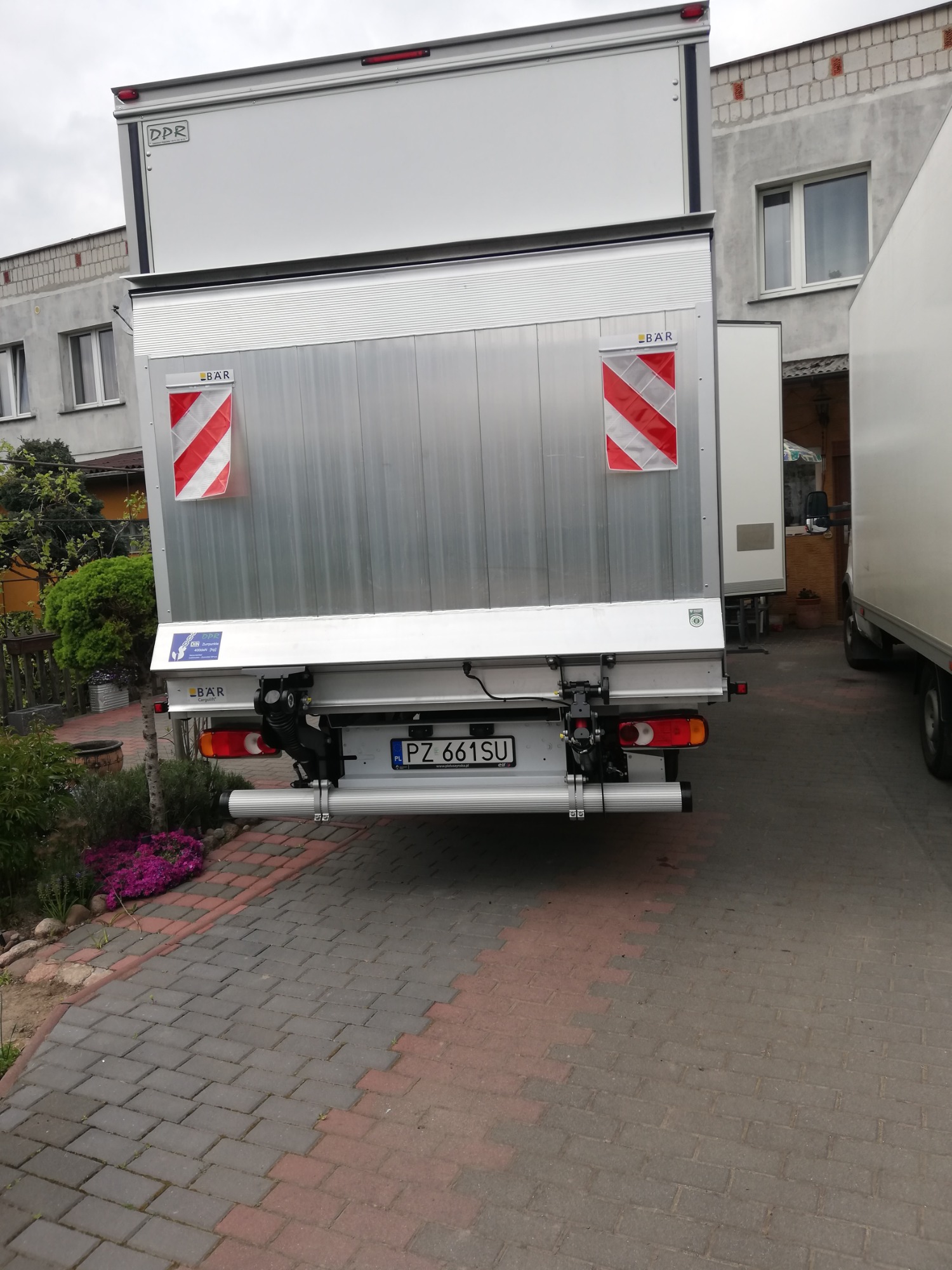 Tył dziewięciopaletowego samochodu dostawczego z windą załadowczą, zaparkowanego na brukowanym podjeździe przed budynkiem mieszkalnym.