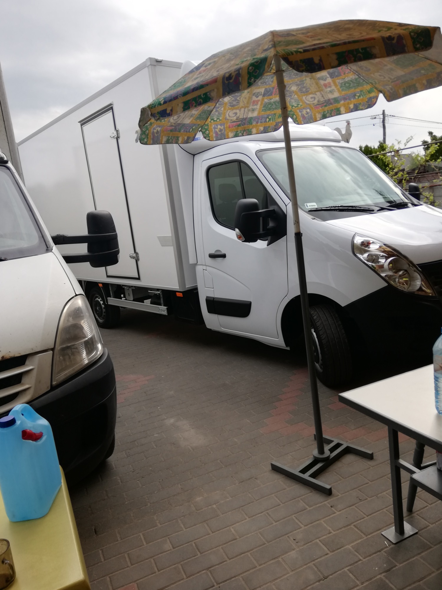 Dwa białe busy dostawcze zaparkowane na brukowanym dziedzińcu, jeden z nadbudową typu kontener, a drugi z widoczną przednią częścią, obok stolika z parasolem przeciwsłonecznym.