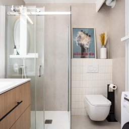 KATARZYNA BENKO OCH-ACH_CONCEPT - Jasna łazienka z przesuwną kabiną prysznicową, umywalką z szafką z drewnianym fornirem, wiszącą toaletą i pralką Beko, uzupełniona plakatem 'Sia la la' i wazonem z suszonymi kwiatami.