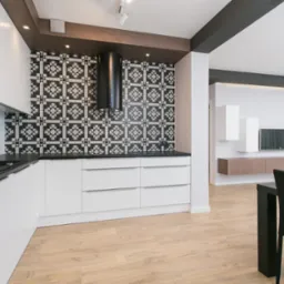 Nowoczesna kuchnia z białymi szafkami, czarnym blatem i wzorzystą czarno-białą ścianą nad blatem, widoczny również salon z telewizorem i szafkami wiszącymi, drewniana podłoga