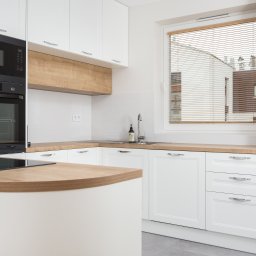 KATARZYNA BENKO OCH-ACH_CONCEPT - Jasna kuchnia z białymi szafkami, drewnianym blatem i zabudowanym piekarnikiem Samsung oraz płytą indukcyjną na wyspie kuchennej, widok na okno z drewnianymi żaluzjami.