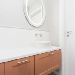 Nowoczesna łazienka z okrągłym lustrem podświetlanym LED, minimalistyczną umywalką nablatową i brązowymi szafkami pod umywalką z chromowanymi uchwytami.