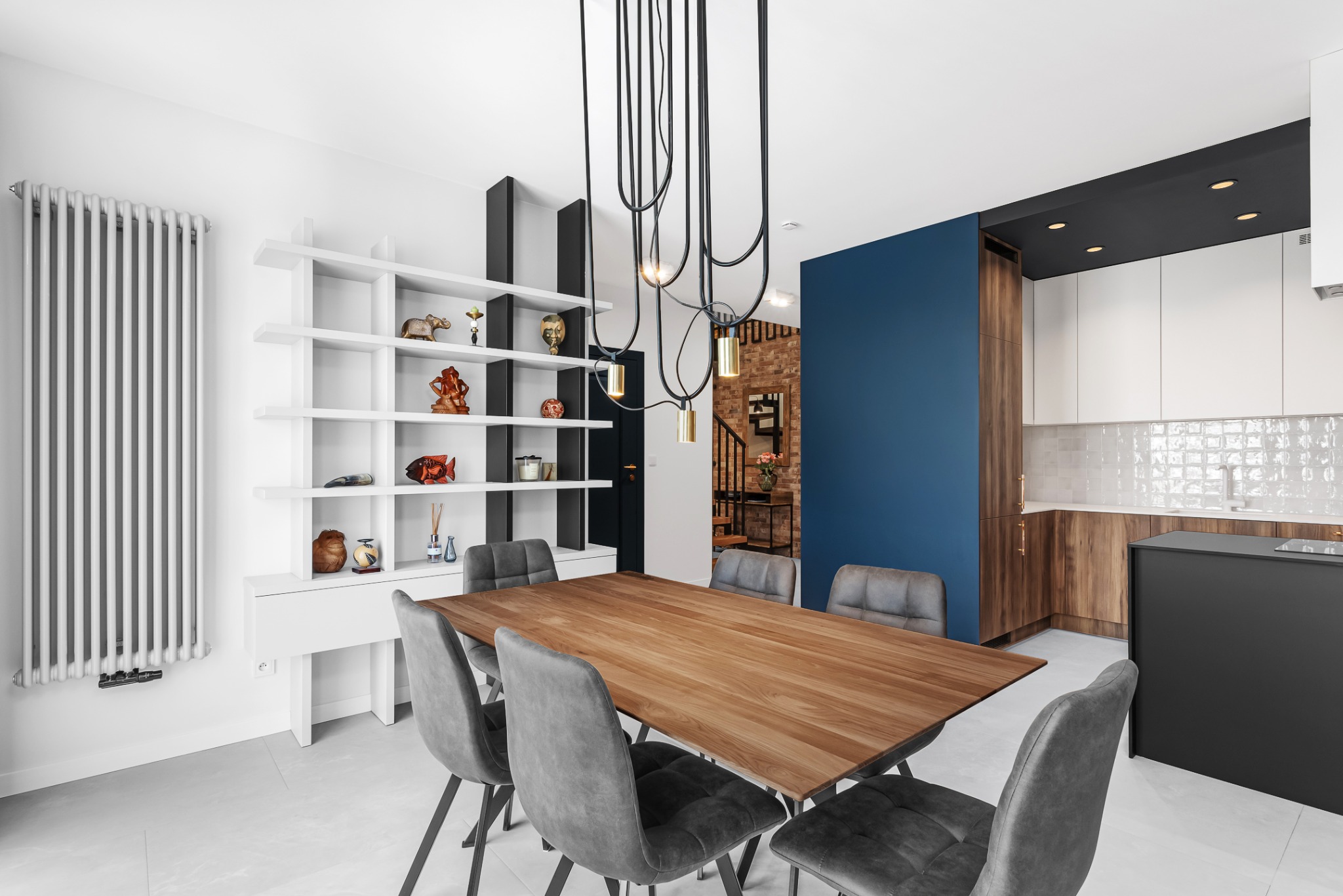 Jasne wnętrze jadalni w Krakowie z drewnianym stołem otoczonym szarymi krzesłami, na tle niebieskiej ściany i otwartej kuchni z białymi szafkami oraz półkami z dekoracjami.