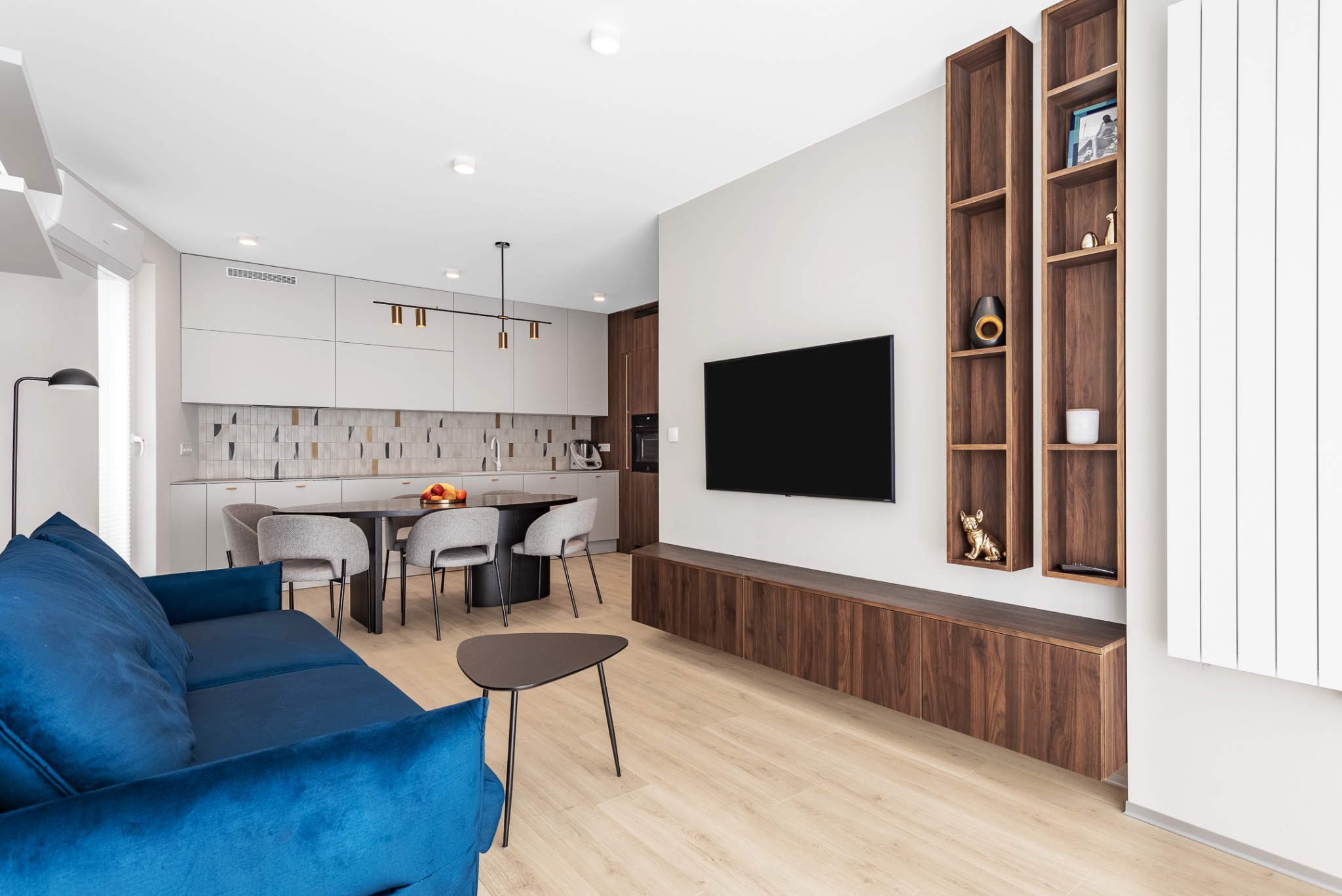 Nowoczesny salon z otwartą kuchnią: niebieska sofa, drewniane regały ścienne, stół z szarymi krzesłami i dekoracyjna mozaika nad blatem kuchennym.