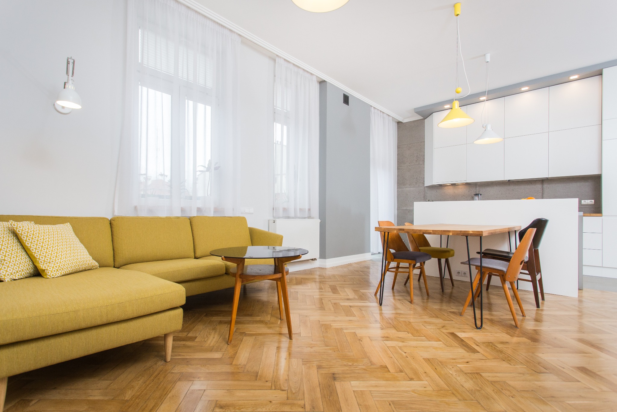 Salon z żółtą sofą narożną i stolikiem kawowym, parkiet w jodełkę, stół jadalny z krzesłami oraz biała zabudowa kuchenna w tle.