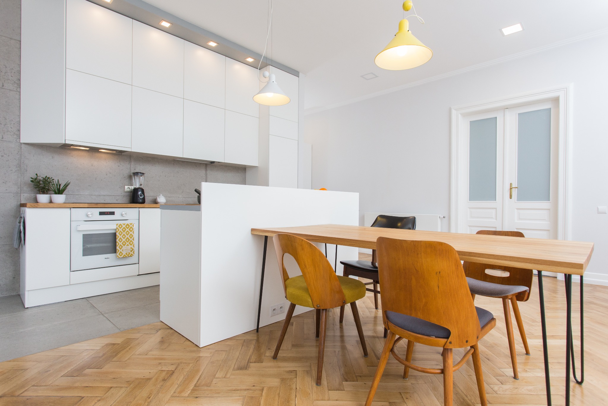 Jasna kuchnia z drewnianym parkietem w jodełkę, białe szafki, drewniany blat, stół z drewnianym blatem i krzesłami w stylu vintage z żółtymi i szarymi siedziskami, wiszące lampy z żółtym kloszem.