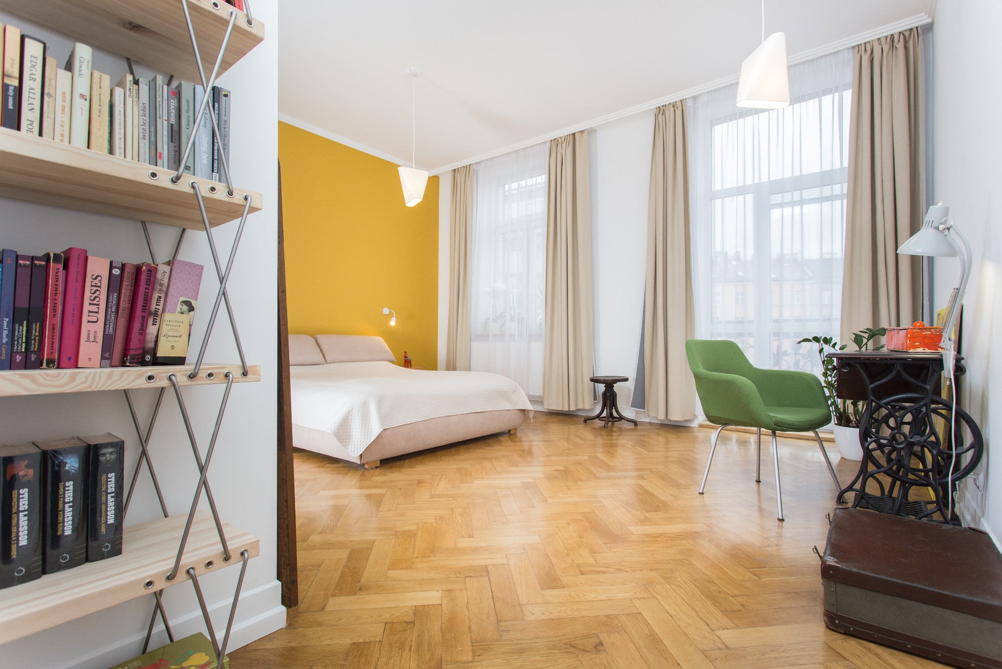 Jasne wnętrze sypialni z żółtą ścianą, widok z salonu z regałem na książki, parkiet w jodełkę, zielone krzesło i stolik z maszyną do szycia.