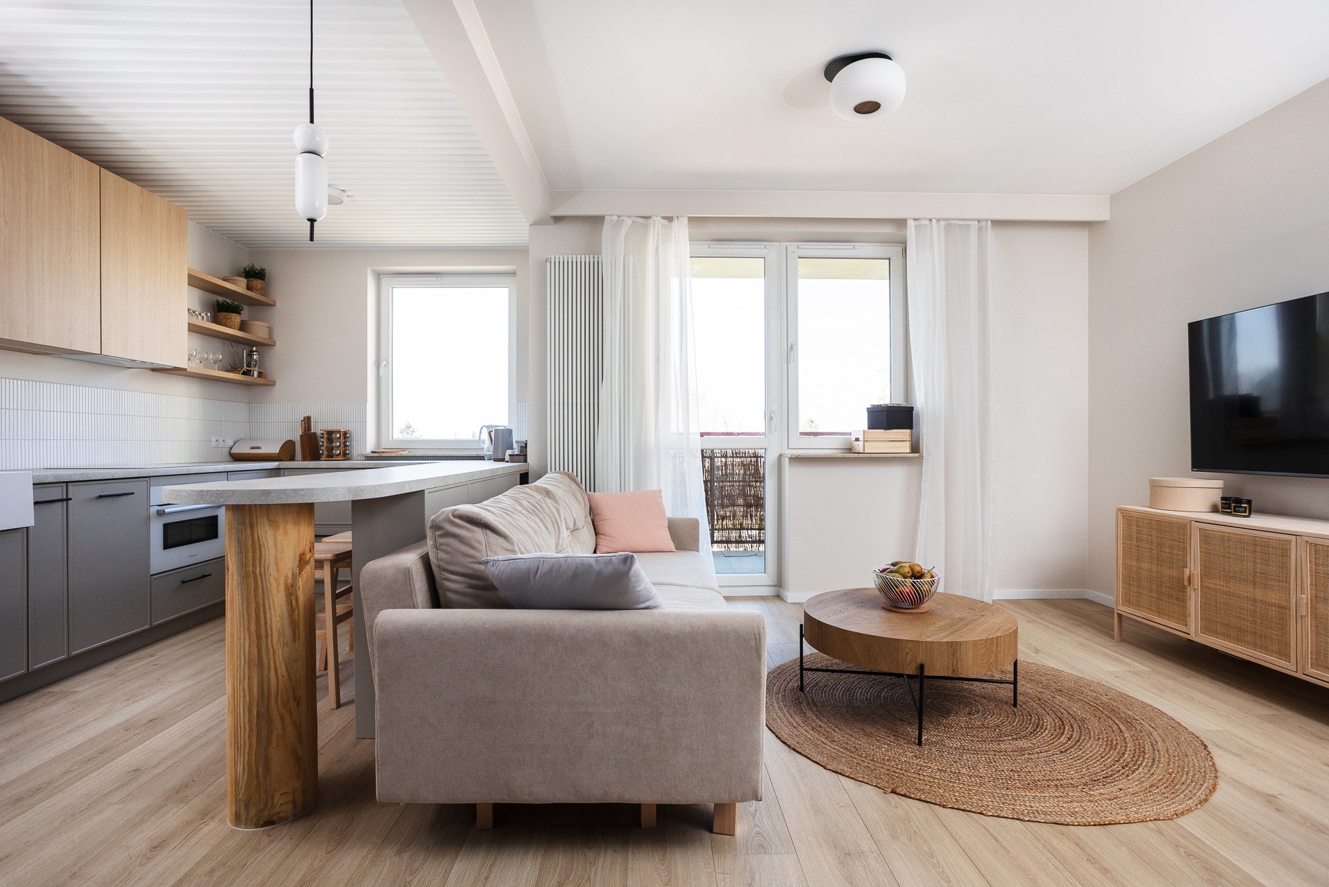 Jasne, nowocześnie urządzone wnętrze mieszkania z aneksem kuchennym, drewnianym blatem wspartym na pniu, beżową sofą z poduszkami i okrągłym dywanem jutowym.