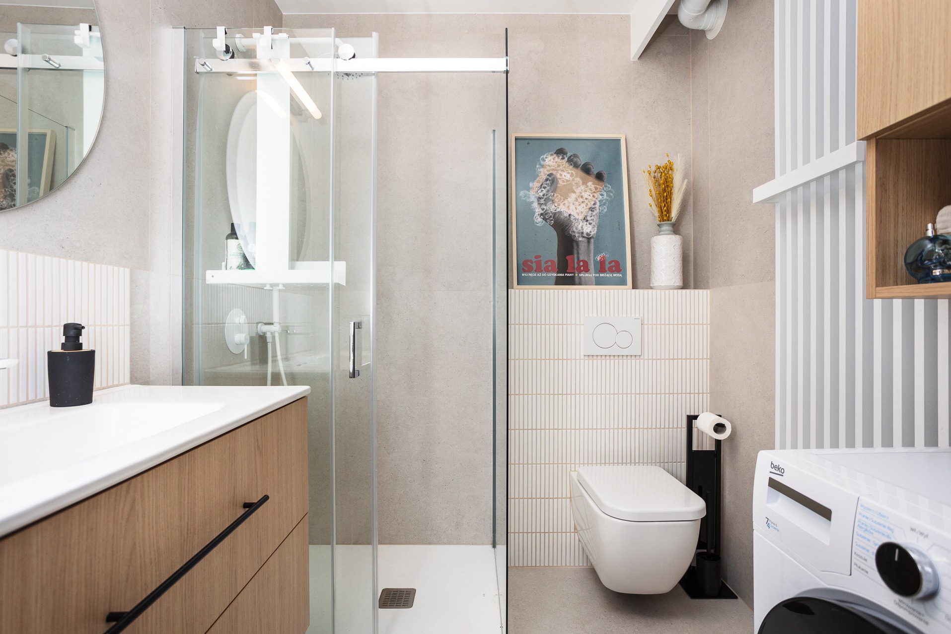Jasna łazienka z przesuwną kabiną prysznicową, umywalką z szafką z drewnianym fornirem, wiszącą toaletą i pralką Beko, uzupełniona plakatem 'Sia la la' i wazonem z suszonymi kwiatami.