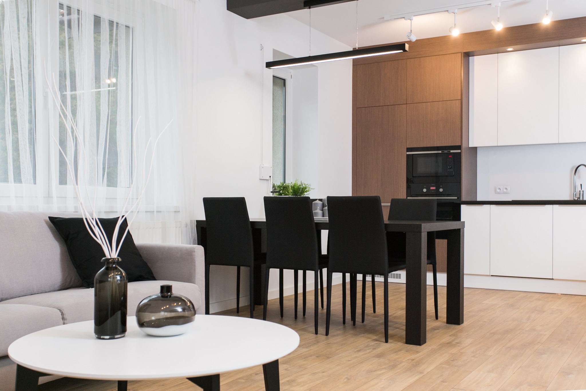 Nowoczesne wnętrze: jasny salon z szarą sofą, okrągłym białym stolikiem kawowym, czarnym stołem jadalnym i krzesłami, oraz kuchnią z drewnianymi i białymi szafkami, oświetlone listwą LED...