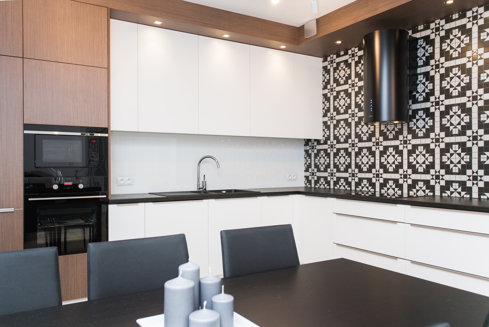 Nowoczesna kuchnia z białymi szafkami, drewnianymi akcentami i czarno-białą mozaiką na ścianie, uzupełniona czarnym okapem cylindrycznym, widok z perspektywy stołu z szarymi świecami.