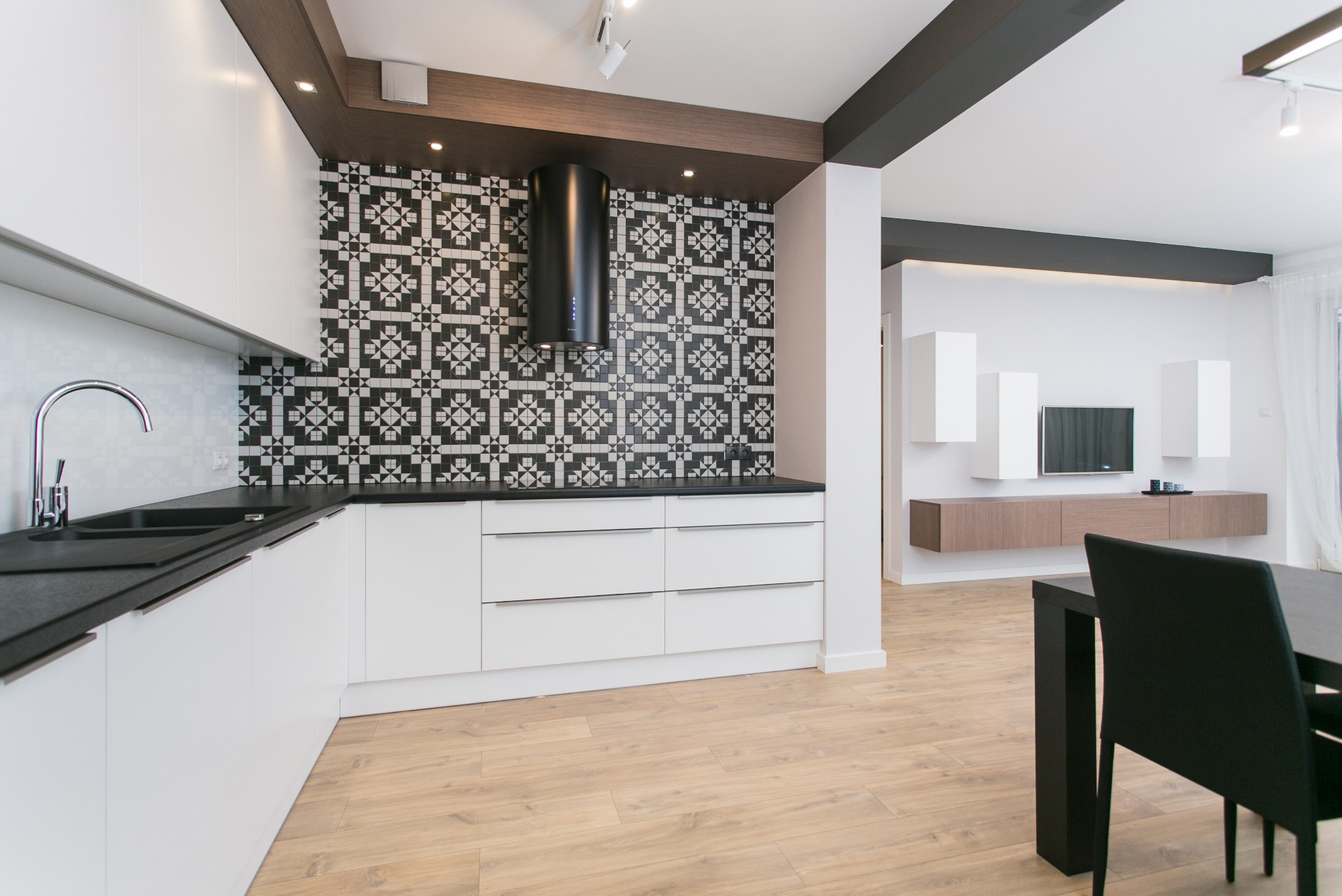 Nowoczesna kuchnia z białymi szafkami, czarnym blatem i wzorzystą czarno-białą ścianą nad blatem, widoczny również salon z telewizorem i szafkami wiszącymi, drewniana podłoga