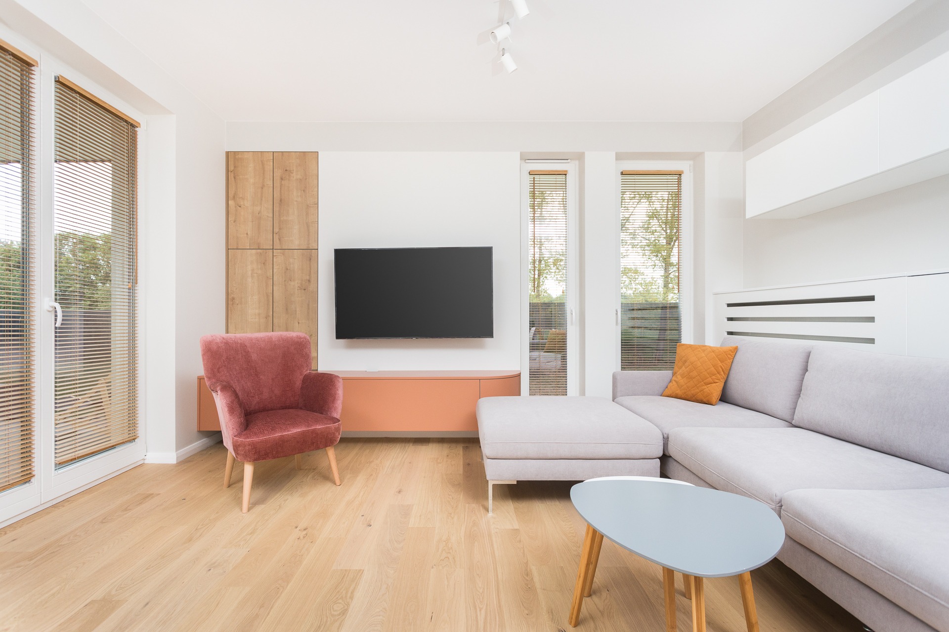 Jasne wnętrze salonu z drewnianą podłogą, szarą sofą narożną z pomarańczową poduszką, różowym fotelem welurowym, stolikiem kawowym w kształcie nerki i telewizorem na ścianie, oświetlonym przez trzy...
