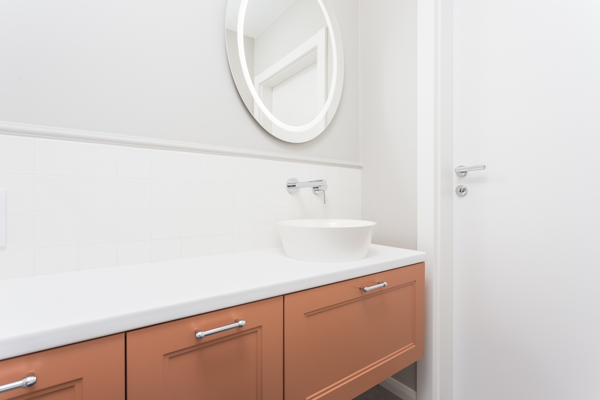 Nowoczesna łazienka z okrągłym lustrem podświetlanym LED, minimalistyczną umywalką nablatową i brązowymi szafkami pod umywalką z chromowanymi uchwytami.