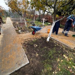 BK Remonty i Serwis - Pracownik w kombinezonie zamiata piasek z nowo ułożonej kostki brukowej na ścieżce ogrodowej, niebieska taczka i wiadro obok drzewa z bielonym pniem.