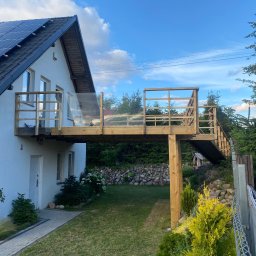 BK Remonty i Serwis - Nowoczesny dom z drewnianym tarasem na piętrze, schody prowadzące do ogrodu, panele słoneczne na dachu, kostka brukowa przed wejściem.