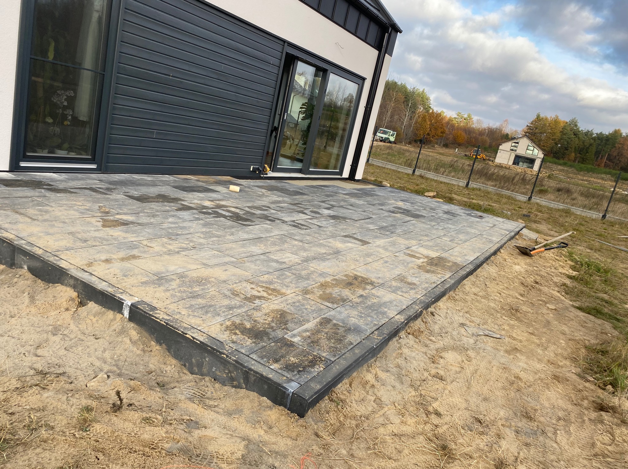 Nowoczesny taras z szarej kostki brukowej w trakcie budowy, widoczne narzędzia pracy i piasek, dom z dużymi oknami w tle.