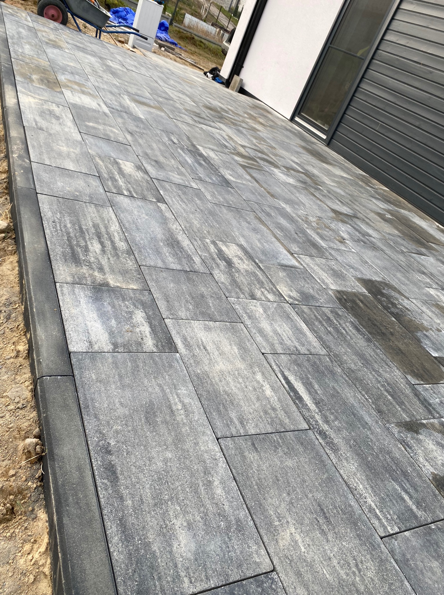 Ułożona szara kostka brukowa z obrzeżami przy nowoczesnym domu z szarą bramą garażową, w tle widoczny wózek budowlany i materiały budowlane.