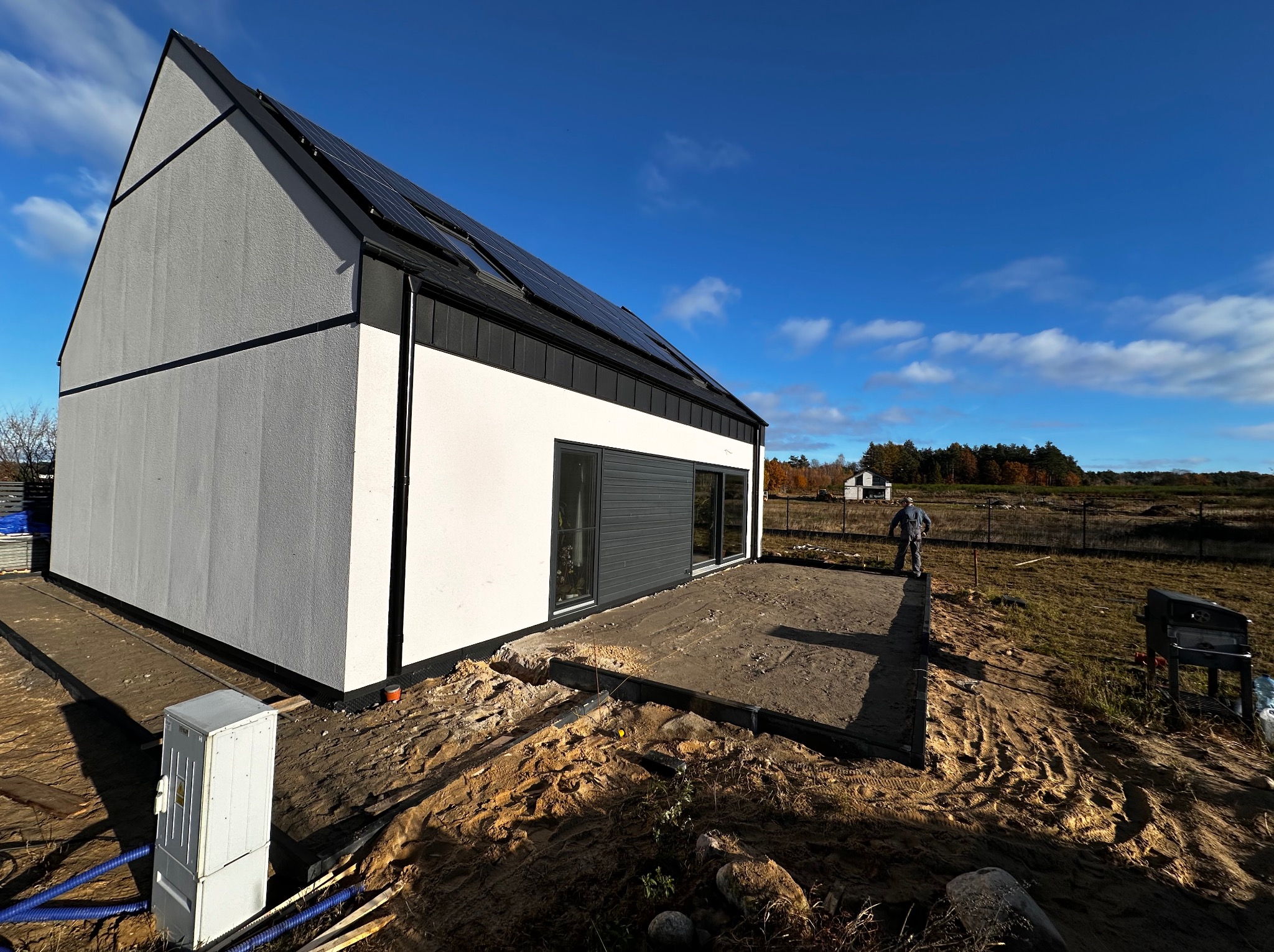 Nowoczesny dom jednorodzinny z panelami słonecznymi w trakcie budowy, widoczny przygotowany teren pod taras i instalacje elektryczne, pracownik w oddali.