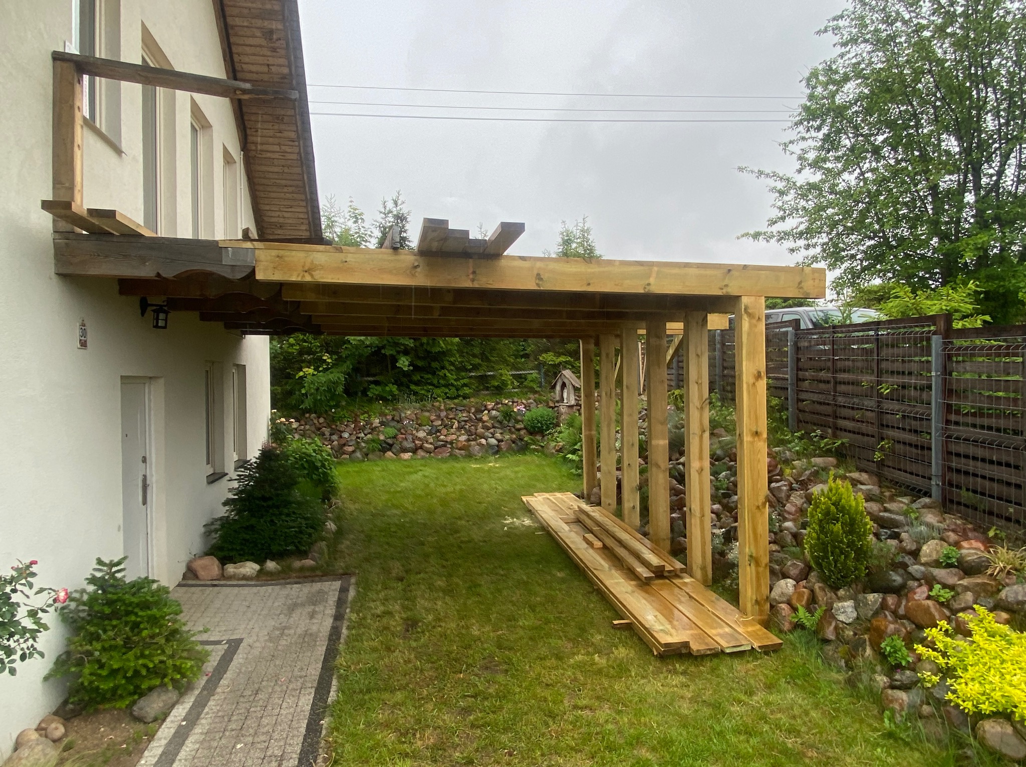 Budowa drewnianej wiaty przy domu: widoczne drewniane słupy, belki konstrukcyjne, przygotowane deski, trawnik, kamienny murek i fragment kostki brukowej.