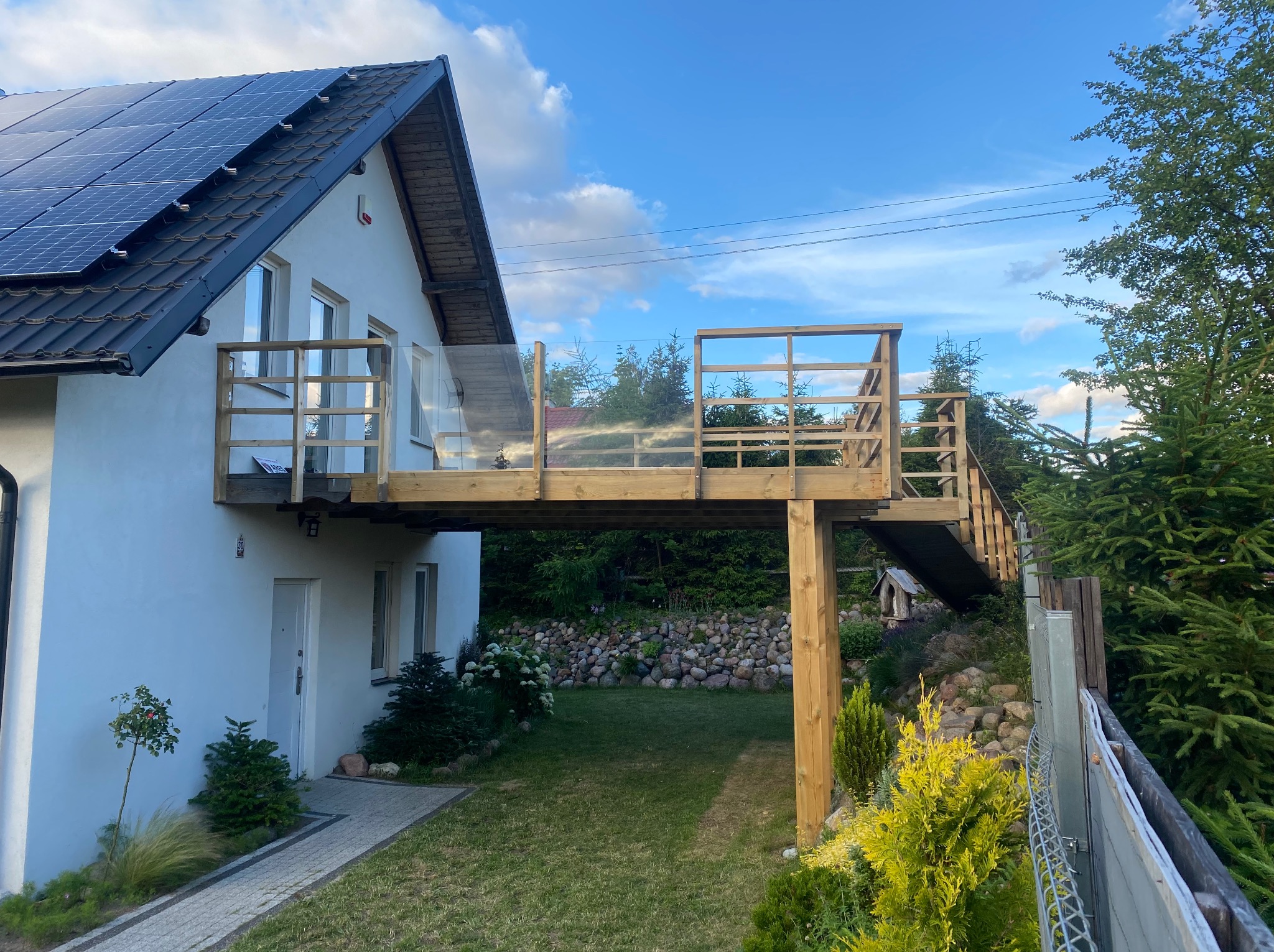 Nowoczesny dom z drewnianym tarasem na piętrze, schody prowadzące do ogrodu, panele słoneczne na dachu, kostka brukowa przed wejściem.