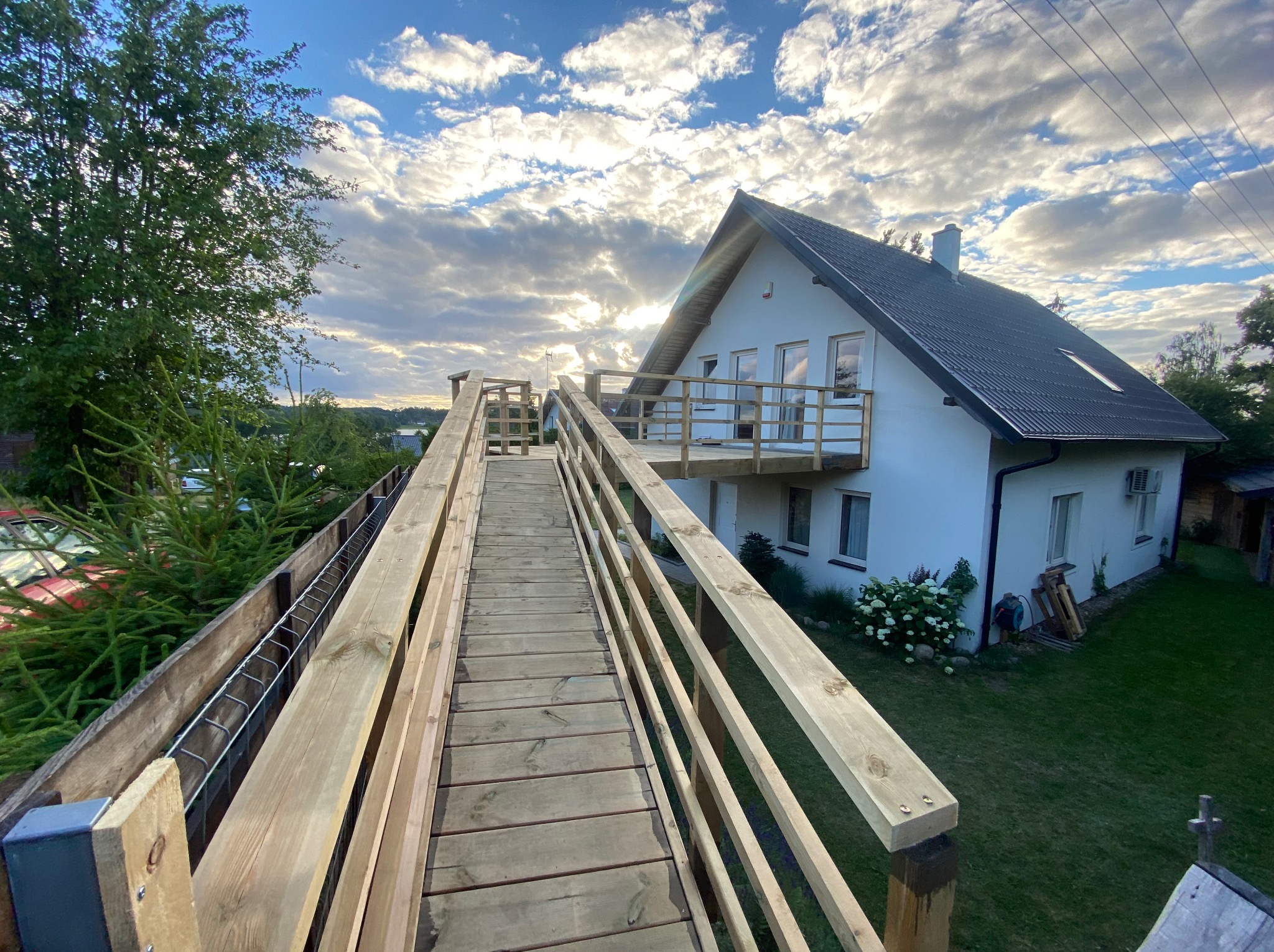 Drewniana kładka dla pieszych prowadząca do domu w Sylcznie, widok z góry na konstrukcję z surowego drewna, w tle biały dom z balkonem i zielony trawnik.
