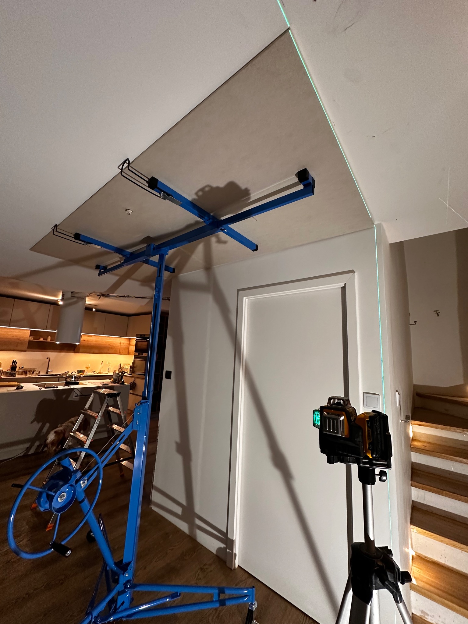 Montaż płyty gipsowo-kartonowej na suficie za pomocą podnośnika, widoczny laser krzyżowy wyznaczający poziom, w tle kuchnia i schody.