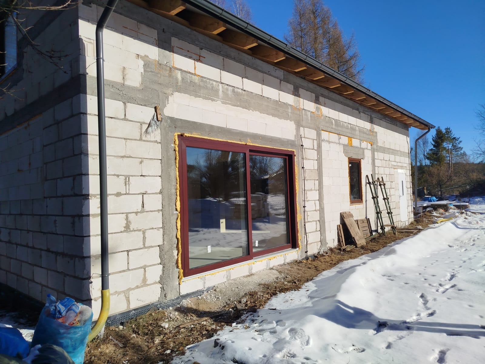 Budynek w trakcie budowy z widocznymi oknami z brązowymi ramami, uszczelnionymi pianką montażową, ściany z białego bloczka, ośnieżony teren, drabina oparta o ścianę, widoczne elementy instalacji...