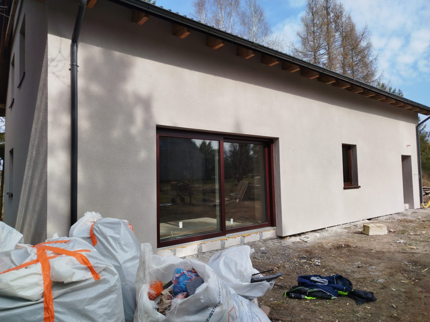 Wykończona elewacja budynku jednorodzinnego z brązowymi oknami, widoczne worki z materiałami budowlanymi i odzieżą roboczą w nieuporządkowanym otoczeniu.