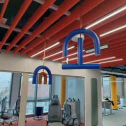 Biuro z nowoczesnym oświetleniem sufitowym w postaci kolorowych, geometrycznych lamp wiszących i listew LED, widok przez szklaną ścianę na pomieszczenie z meblami biurowymi w folii ochronnej.