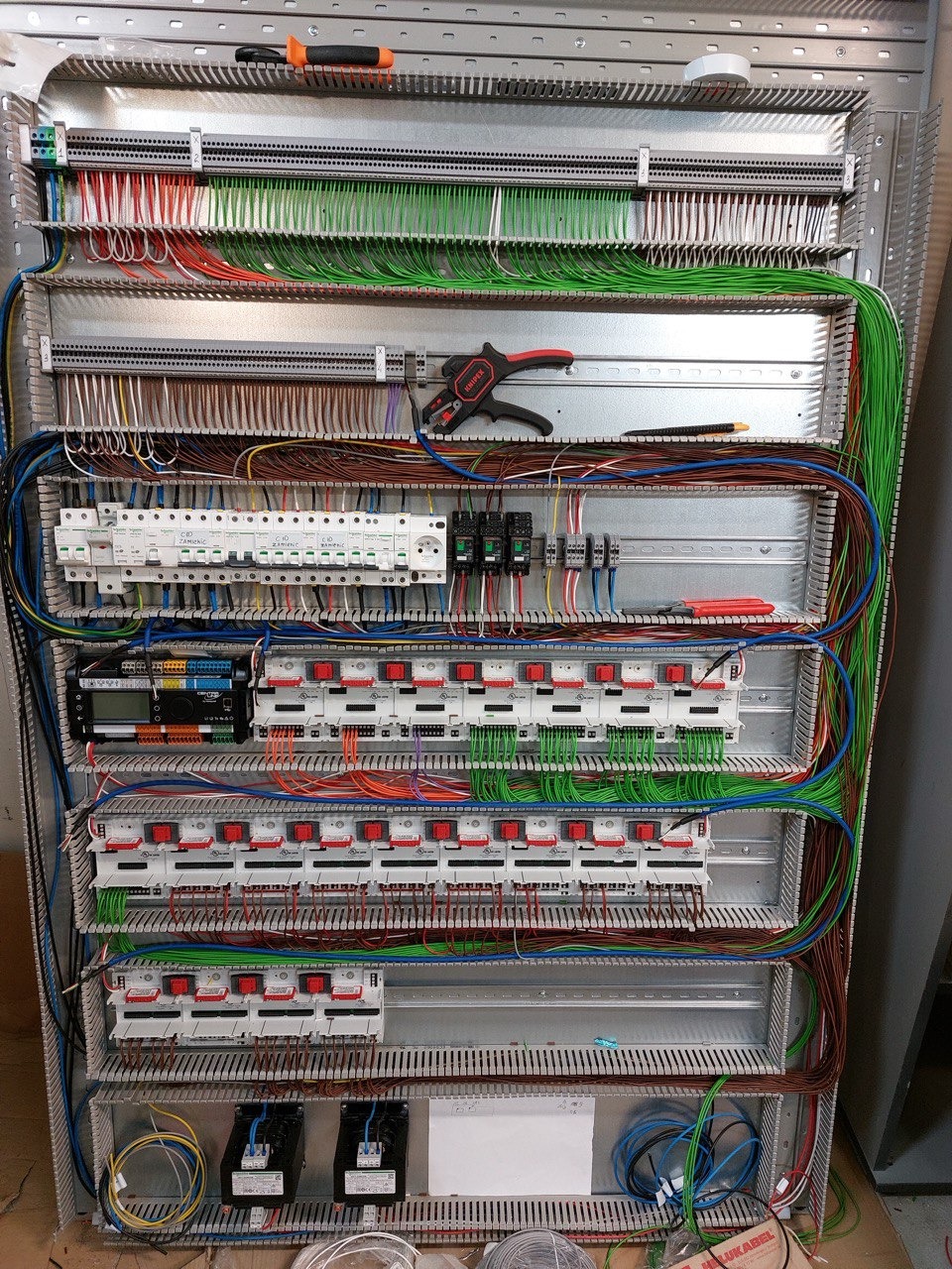 Rozdzielnica elektryczna w trakcie montażu, widoczne liczne przewody, zabezpieczenia i narzędzia: nóż do ściągania izolacji i kombinerki.