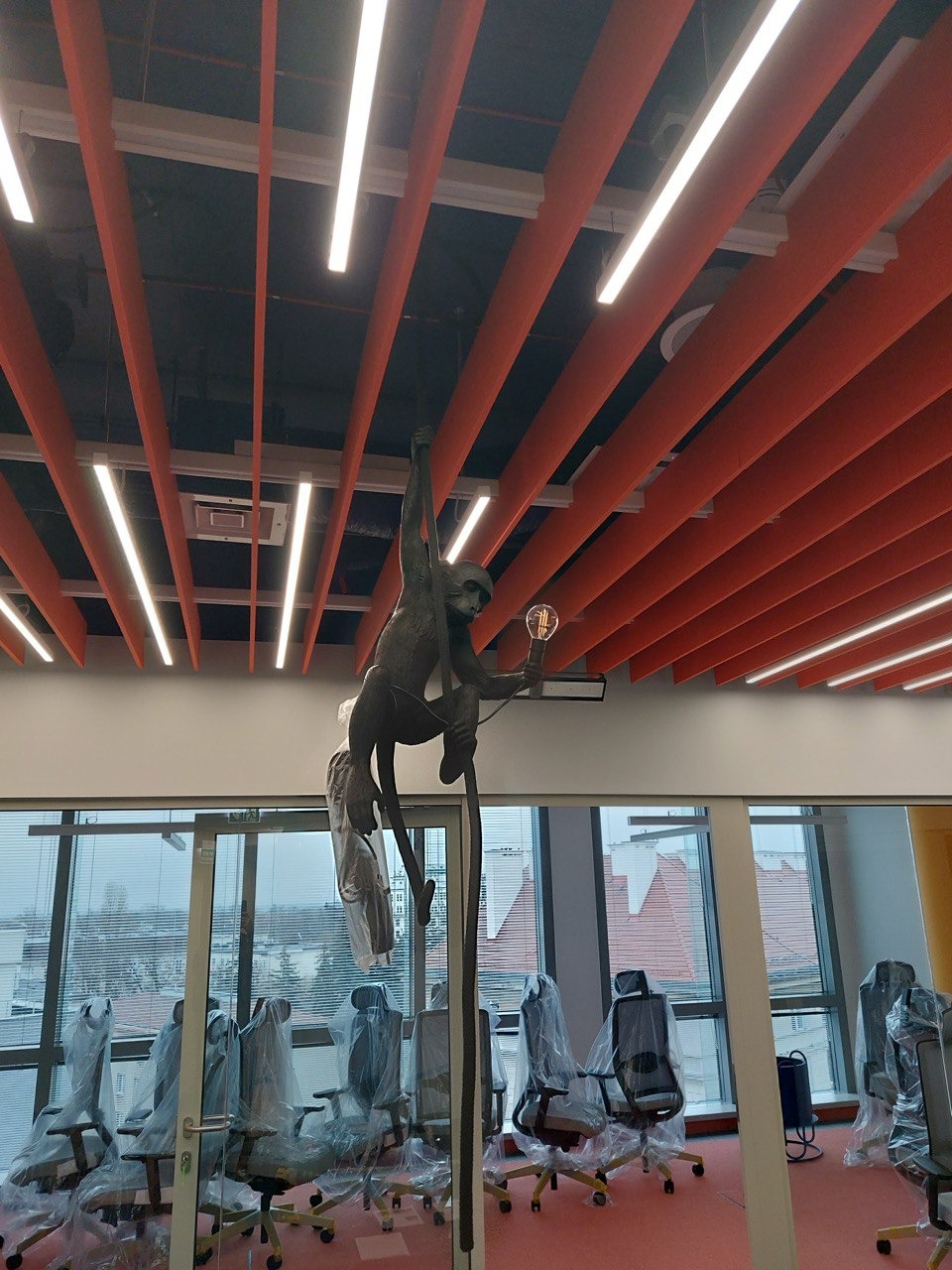Wnętrze biurowe z rzeźbą małpy trzymającej żarówkę, widoczne lampy sufitowe, krzesła biurowe w folii ochronnej oraz okna z widokiem na miasto.