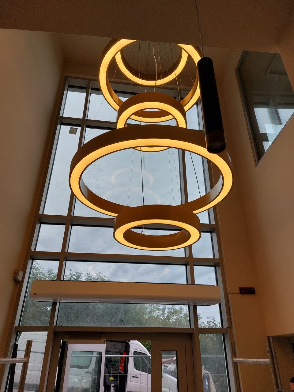 Nowoczesna, okrągła lampa wisząca z czterema pierścieniami LED, zamontowana w wysokim holu z dużym oknem i widocznym samochodem dostawczym na zewnątrz.