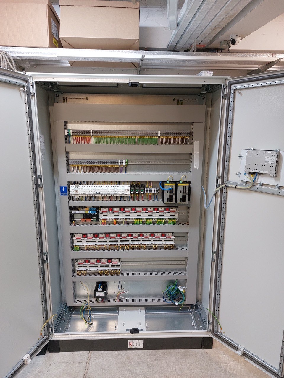 Otwarta szafa sterownicza z widocznymi elementami instalacji elektrycznej, w tle sufit z instalacjami i kartonami.